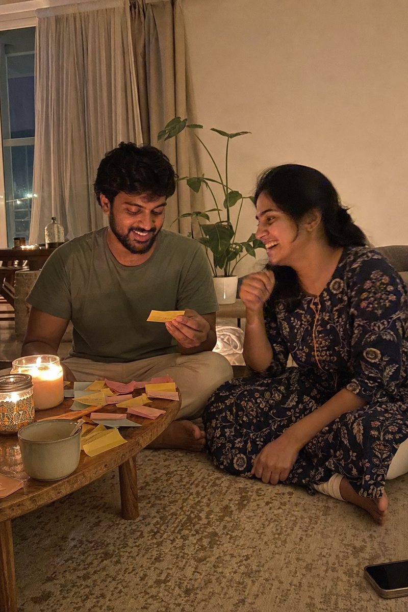 मोमबत्तियों से सजी कॉफ़ी टेबल के पास फ़र्श पर हँसता एक कपल, टेबल पर पेस्टल रंग के स्टिकी नोट बिखरे हैं